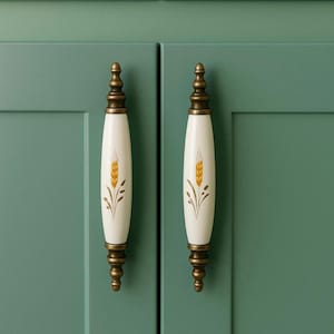 May include: Two cream-colored cabinet pulls with a wheat stalk design. The pulls have a brass-colored base and top, and are mounted on a green cabinet door. The wheat design is in shades of gold and brown.