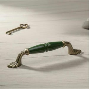 May include: A decorative cabinet pull with a dark green ceramic center and antique brass-colored metal ends. The pull has a curved design and is accompanied by a vintage key.