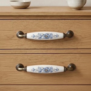 May include: Two wooden cabinet drawers with white ceramic handles featuring a blue floral design and antique brass fittings. The drawers are made of light-coloured wood. The handles are approximately 15 cm long.
