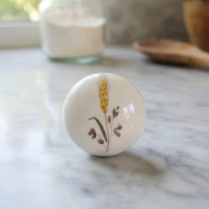 May include: A white ceramic cabinet knob with a wheat stalk design. The wheat is yellow, with brown and gray accents. The knob is round and glossy, suitable for kitchen or furniture decor.