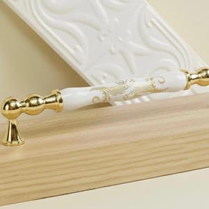 Può includere: Una maniglia per mobili bianca e dorata con un motivo floreale. La maniglia ha un manico in ceramica bianca con dettagli dorati e una base dorata. La maniglia è lunga circa 15 cm.