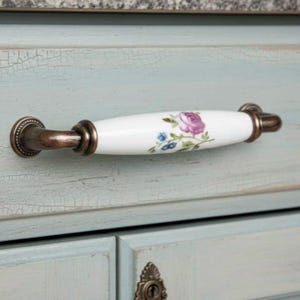 Puede incluir: Un tirador de armario con un mango de cerámica blanca con un diseño floral de rosa púrpura y azul. Los extremos son de metal color bronce. El tirador está montado en una puerta de armario azul claro.