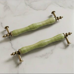 May include: Two cabinet pulls with a light green, textured handle and antique brass hardware. The handles have a cylindrical shape with a slightly curved design. The brass hardware includes decorative elements and mounting posts.