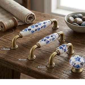 May include: Cabinet hardware set featuring handles and a knob. The handles have white ceramic centres adorned with blue floral patterns and brass-toned metal bases. The largest handle is 12.8cm long. The knob has a matching floral design.