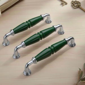 Peut inclure: Trois poignées de meubles avec un centre cylindrique vert foncé et des extrémités incurvées argentées. Les poignées sont disposées sur une surface en bois de couleur claire. Quincaillerie décorative pour meubles.