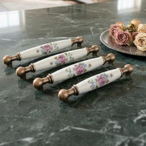 May include: Four white ceramic cabinet pulls with bronze-colored hardware. Each pull features a floral design with pink roses and blue accents. The pulls are arranged on a green marble surface.