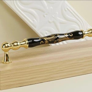 May include: A decorative cabinet pull with a black center and gold accents. The pull features a gold floral design and gold-colored ends. The pull is mounted on a light-colored wooden surface.