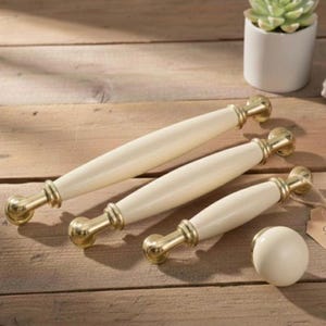Op de afbeelding: Crèmekleurige kastgrepen en een bijpassende knop met goudkleurige hardware. De grepen zijn ovaal van vorm en variëren in grootte. De knop is rond. De hardware heeft een warme goudkleur. Op een houten oppervlak.