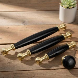 Op de afbeelding: Vier zwarte en gouden kastgrepen en een knop liggen op een houten oppervlak. De grepen hebben een slank, langwerpig ontwerp met zwarte centra en gouden accenten. De knop is rond en zwart.