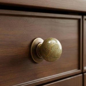 Op de afbeelding: Close-up van een houten lade met een decoratieve knop. De knop is een bol met een gemarmerd groen en goud patroon, geplaatst op een cirkelvormige, antieke messing basis. De lade is gemaakt van donkerbruin hout.