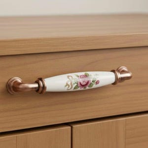 Pode incluir: Um puxador de armário com uma pega de cerâmica branca com um desenho de rosa rosa. A pega é acentuada com ferragens metálicas de cor bronze. O puxador é montado num armário de madeira castanha clara.