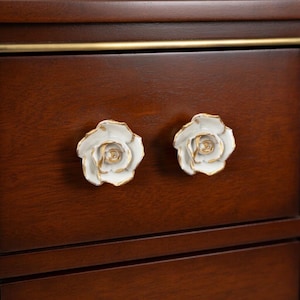 Può includere: Primo piano di un cassetto in legno con due pomelli a forma di rosa bianca. I pomelli hanno dettagli color oro e sono montati su una superficie in legno marrone scuro. Il cassetto ha un bordo dorato.