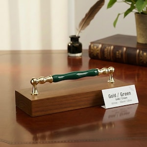 Op de afbeelding: Een goud-groene kastgreep. De greep heeft een donkergroen midden en goudkleurige uiteinden. De greep wordt op een houten blok gepresenteerd. Het label luidt "Gold / Green".