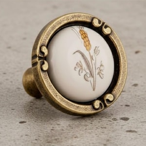 Könnte beinhalten: Antiker Messing-Schrankknauf mit weißem Keramikmittelteil mit Weizenähren-Design. Der Knauf hat eine runde Form mit dekorativen Details und einem kleinen, abgerundeten Griff.