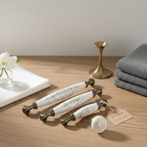 Könnte beinhalten: Ein Set aus vier weißen und bronzefarbenen Schrankgriffen und einem passenden Knauf mit floralem Design. Die Griffe sind in verschiedenen Größen erhältlich und auf einer hellen Holzoberfläche angeordnet. Ein kleines Etikett ist am Knauf befestigt.