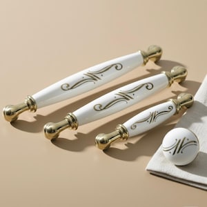 Puede incluir: Un juego de herrajes para armarios blancos y dorados, que incluye tres tiradores y un pomo. Los tiradores tienen un cuerpo de cerámica blanca con detalles dorados y remolinos dorados decorativos. El pomo es una esfera blanca con un diseño de remolino dorado.