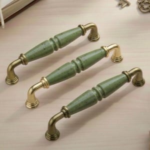 May include: Three cabinet handles with a gold finish and speckled green ceramic centres. The handles are arranged diagonally on a light wood surface. The hardware has a classic, elegant design.