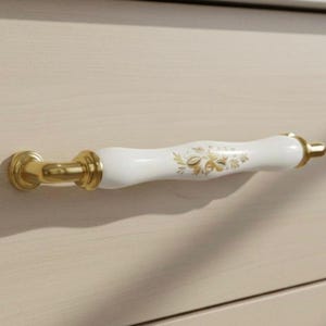 Può includere: Una maniglia per armadio bianca e dorata con un design floreale. La maniglia è bianca con dettagli dorati e un motivo floreale dorato. L'hardware di montaggio è dorato. L'anta dell'armadio è di colore beige chiaro.