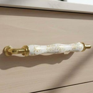 May include: A cabinet pull featuring a white and gold speckled ceramic handle and gold-toned metal fixtures. The handle is attached to a light-coloured wooden cabinet.