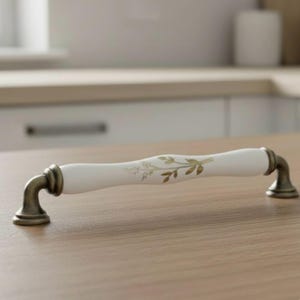 Puede incluir: Tirador de armario de cerámica blanca con diseño floral dorado. El tirador tiene forma curvada con detalles metálicos color bronce en los extremos. Aproximadamente 15 cm de largo, ideal para armarios de cocina o baño.