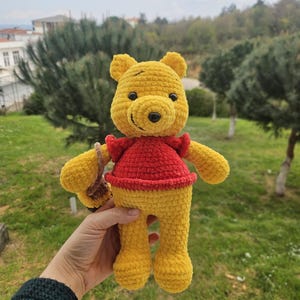 Könnte beinhalten: Ein handgefertigtes gelbes Winnie-Puuh-Plüschtier mit rotem Hemd und kleinem braunen Korb. Das Plüschtier besteht aus einem weichen, strukturierten Material und hat schwarze Augen und eine schwarze Nase. Das Spielzeug ist etwa 25 cm groß.