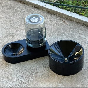 Puede incluir: Un juego de cuencos de agua y comida para mascotas de madera negra con un recipiente de agua de vidrio transparente. El juego incluye dos cuencos y un recipiente de agua. Los cuencos tienen un acabado brillante y un diseño circular.