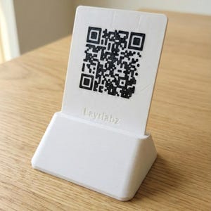 May include: A white 3D-printed stand displaying a QR code. The black QR code is a square with a detailed pattern. The stand has a rectangular base and a sloping top. The text "Layrlabz" is printed beneath the QR code.