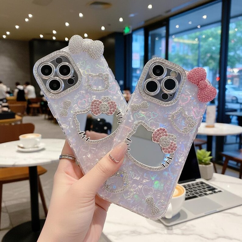 May include: Two iridescent phone cases with a Hello Kitty design. Each case features a bow and a decorative border of rhinestones around the camera and the Hello Kitty face. The cases have a clear, sparkly finish.