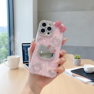 May include: Pink phone case with a Hello Kitty design. The case features a mirror, a bow, and rhinestone accents. The camera lens is surrounded by rhinestones. The case is held in a person's hand.
