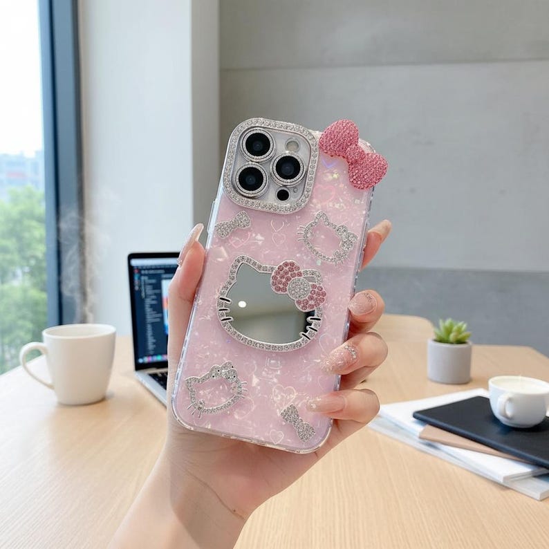 May include: Pink phone case with a Hello Kitty design. The case features a mirror, a bow, and rhinestone accents. The camera lens is surrounded by rhinestones. The case is held in a person's hand.