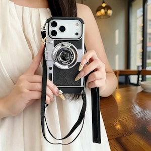 Puede incluir: Funda de teléfono negra y plateada diseñada para parecerse a una cámara vintage. La funda tiene una correa negra y un detalle de lente plateada con texto. La funda del teléfono la sostiene una persona.