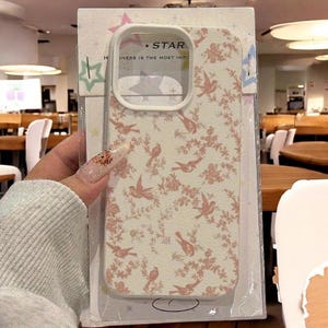 May include: A phone case with a floral and bird pattern in pink and cream. The case has a white border and a cutout for the camera. The text "STAR" and "HAPPINESS IS THE MOST IMPO" are visible on the case.