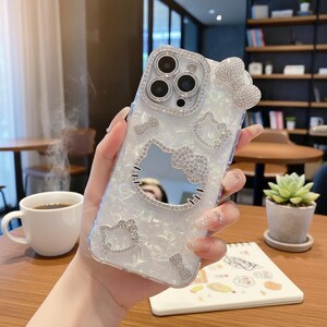 May include: Clear phone case with a pearlescent finish, adorned with sparkling rhinestones in the shape of Hello Kitty and a bow. The case has a mirrored Hello Kitty face cutout and is embellished with additional rhinestone accents.