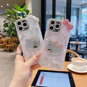 May include: Two iridescent phone cases with Hello Kitty designs. Each case features a bow at the top and a Hello Kitty face with a mirrored finish. The cases are decorated with rhinestones and are held in a person's hand.