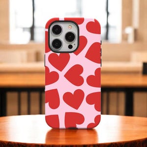 May include: A red and pink phone case with a pattern of red hearts on a pink background. The case has a red border and a camera cutout. The phone case is on a wooden table.