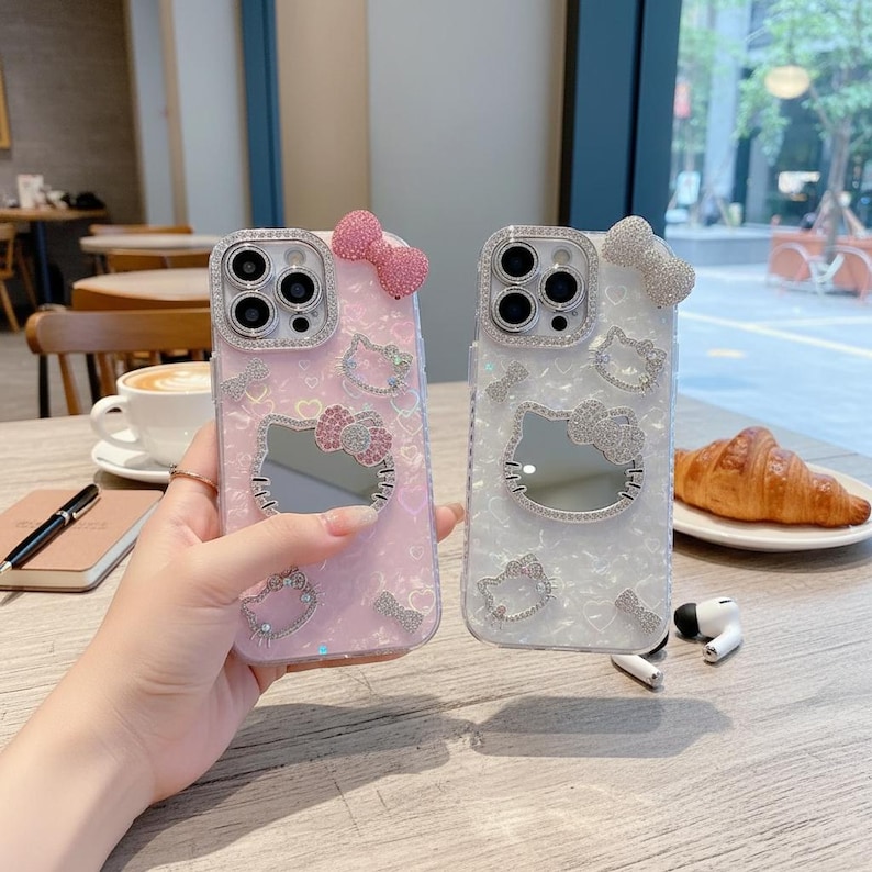 May include: Two phone cases, one pink and one white, featuring a Hello Kitty design. Each case has a mirrored Hello Kitty face, embellished with rhinestones and a bow. The cases are decorated with additional rhinestone accents.