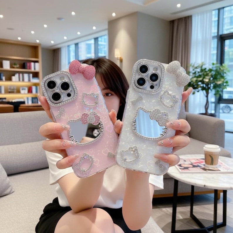 May include: Two phone cases, one pink and one white, each adorned with sparkling embellishments. The cases feature a Hello Kitty design with a bow and a mirror. The pink case has a pink bow, while the white case has a silver bow.