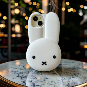 Op de afbeelding: Een witte telefoonhoes in de vorm van een cartoonkonijn met lange oren. De hoes heeft zwarte stipogen en een zwarte 'x' als mond. De cameralenzen van de telefoon zijn zichtbaar in het midden van de hoes.