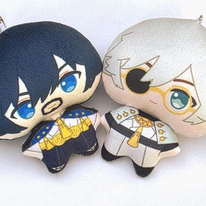 Puede incluir: Dos llaveros de peluche de personajes. Uno tiene cabello azul oscuro y ojos azules, el otro tiene cabello blanco y gafas. Ambos tienen pequeños trajes detallados en azul marino, dorado y blanco. Cada llavero tiene una cadena plateada.
