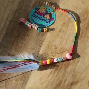 May include: A decorative feather with colorful string accents and a beaded strand spelling "CUTE". A round teal tag with the text "ZauBella" and fairy illustrations is also present. The items are arranged on a wooden surface.