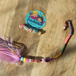 May include: A light pink feather attached to a multi-colored thread cord featuring plastic beads that spell out WILD, a small pink butterfly charm, and several thin metallic threads. A round sticker with the text ZauBella is also visible.