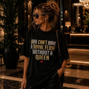 May include: A black short-sleeved t-shirt featuring the gold and white text "YOU CAN'T HAVE A ROYAL FLUSH WITHOUT A QUEEN" printed on the front, accompanied by a small red heart symbol positioned directly below the lettering.