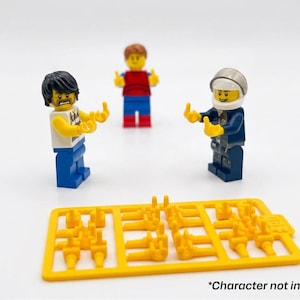 Może przedstawiać: Trzy minifigurki Lego w r&oacute;żnych strojach i z r&oacute;żnymi wyrazami twarzy. Jedna z figurek ma na sobie niebieski mundur i hełm. Ż&oacute;łty plastikowy wlew z kilkoma małymi elementami dłoni znajduje się na pierwszym planie. Tekst na dole brzmi "*Character not included".