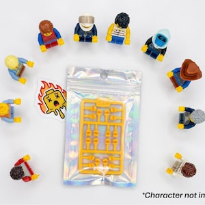 Może przedstawiać: Kolekcja kolorowych figurek Lego ułożonych w okrąg wok&oacute;ł zamkniętej holograficznej torby zawierającej ż&oacute;łte akcesoria Lego. Obecna jest r&oacute;wnież naklejka z płonącą głową Lego. Tekst "*Character not included" znajduje się na dole.