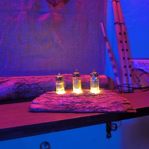 May include: Three small, clear glass vials with gold-colored tops are illuminated with a warm yellow light. They are arranged on a rough, dark gray, rectangular stone slab. The background includes a wooden shelf, a wall, and musical instruments.