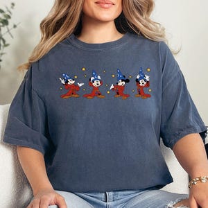 May include: A blue-grey t-shirt featuring an embroidered design of four cartoon characters in red robes and blue wizard hats. The characters are in a row, with their arms raised. The shirt is paired with ripped blue jeans.