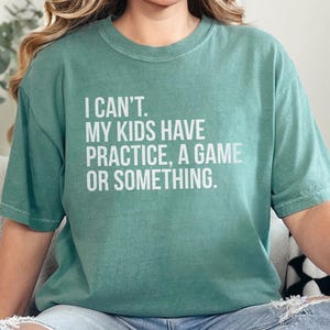 May include: A green short-sleeved t-shirt with the text "I CAN'T. MY KIDS HAVE PRACTICE, A GAME OR SOMETHING." in white. The shirt is worn with blue jeans. The overall theme is casual wear.