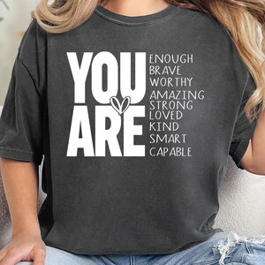 May include: A white ceramic mug with a black handle and the text "But First, Coffee" in a black, handwritten-style font. The mug is on a white surface.