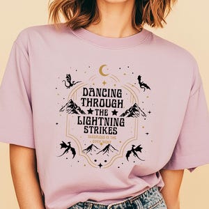 Puede incluir: Camiseta de color lila con un dise&ntilde;o gr&aacute;fico que dice "Dancing Through The Lightning Strikes". El dise&ntilde;o incluye monta&ntilde;as, estrellas y siluetas de dragones. La camiseta tiene mangas cortas y cuello redondo.