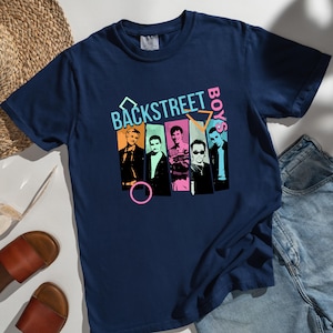 May include: Navy blue t-shirt featuring a graphic of the Backstreet Boys with a retro design. The shirt includes the band's name in a colorful, stylized font. The image is set against a backdrop of denim shorts, sandals, and a straw hat.